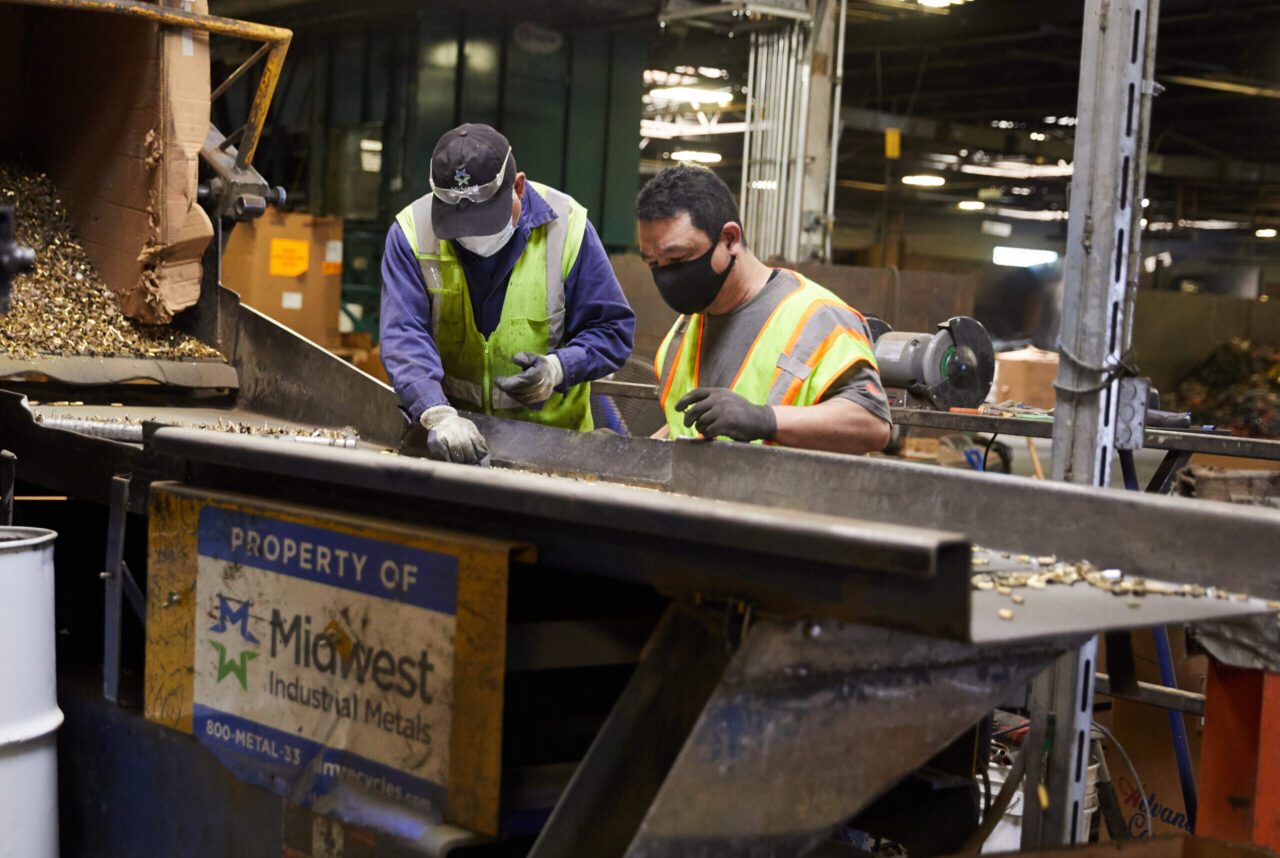 Two men wearing safety vests and masks working on a metal object.